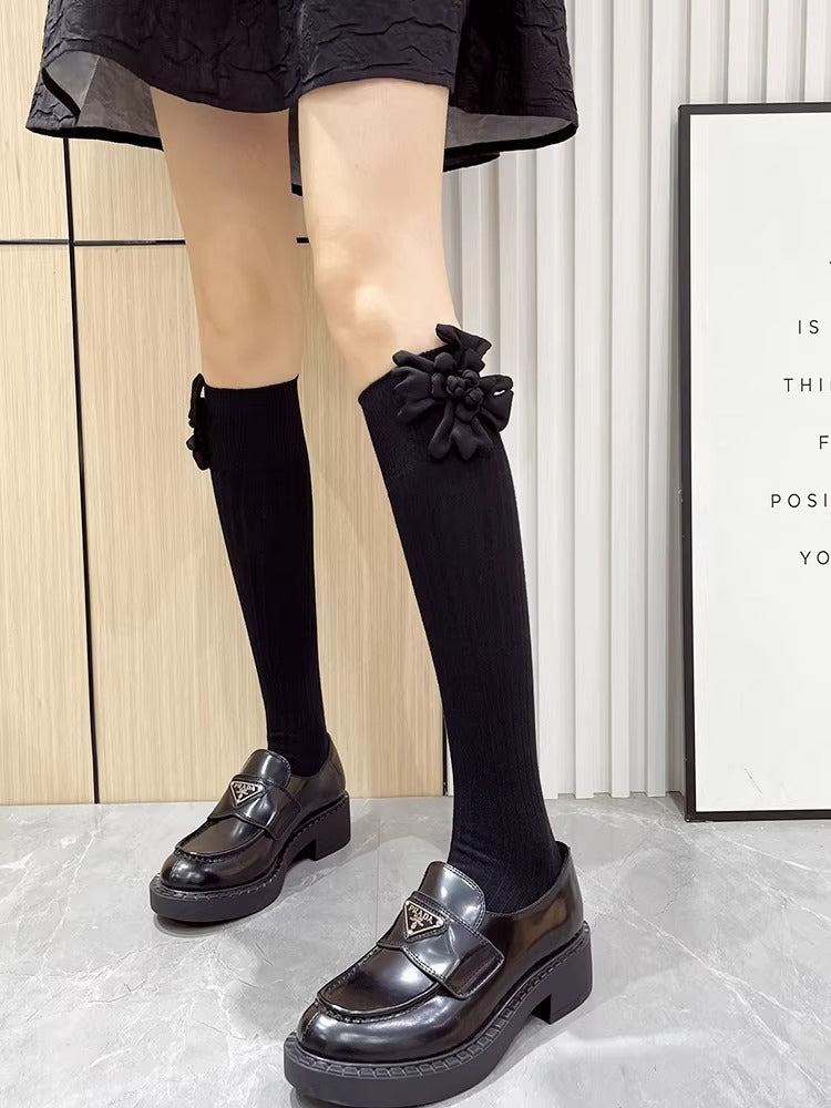person wearing black knee-high ribbed socks with a flower embellishment and black loafers