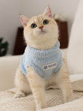 A light blue fluffy knit pet vest with a mock neck and a white logo patch on the front, worn by a light-colored cat.