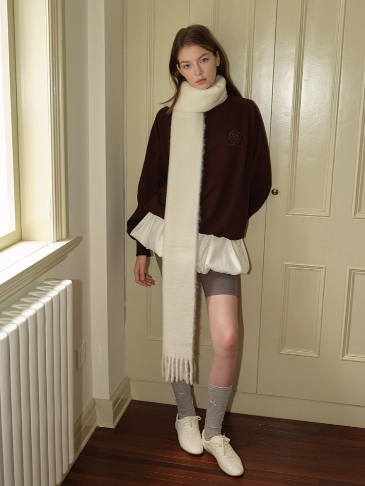 Woman wearing a brown velour sweatshirt with a white scarf, white ruffled undershirt, grey shorts, grey socks, and white sneakers, standing near a door.