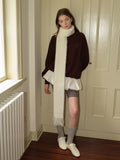 Woman wearing a brown velour sweatshirt with a white scarf, white ruffled undershirt, grey shorts, grey socks, and white sneakers, standing near a door.