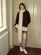 Woman wearing a brown velour sweatshirt with a white scarf, white ruffled undershirt, grey shorts, grey socks, and white sneakers, standing near a door.