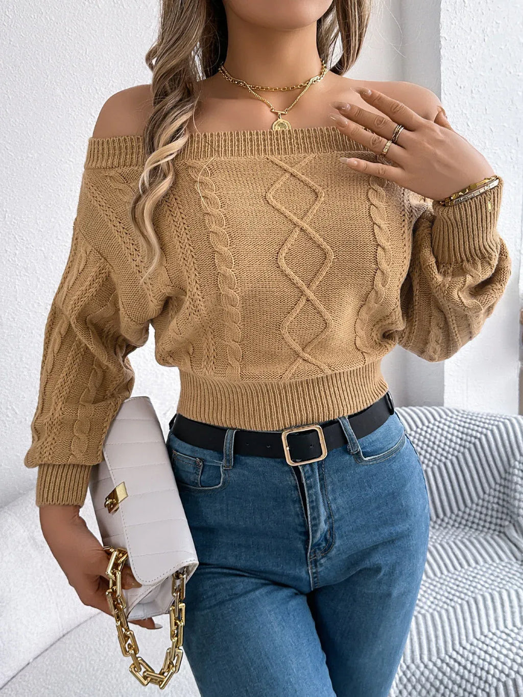 Close-up of a woman wearing a tan off-the-shoulder cable knit cropped sweater, blue jeans, and jewelry.