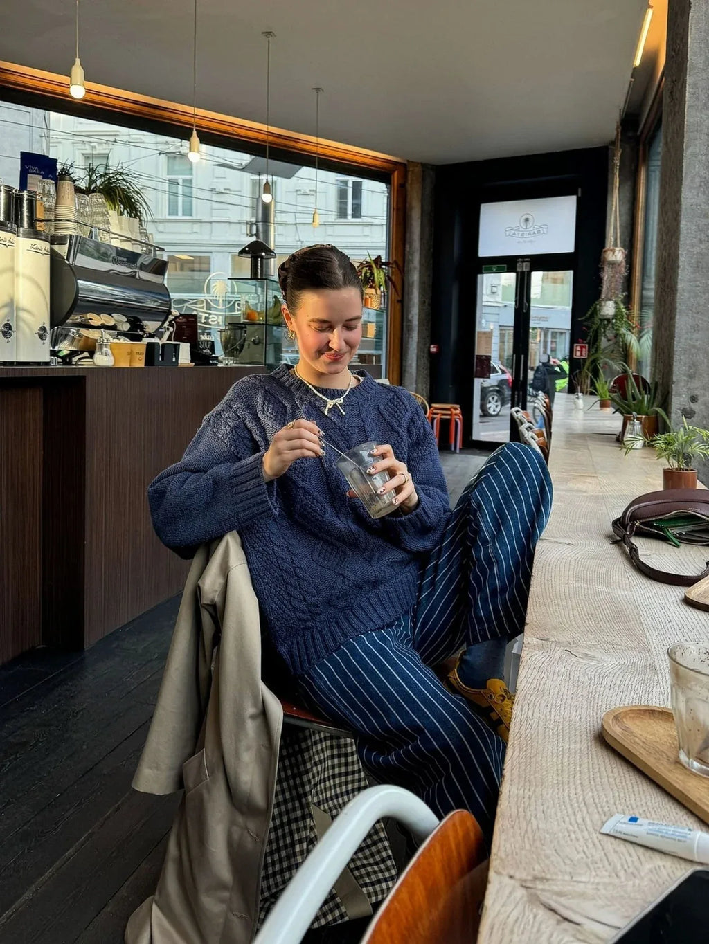 Woman sitting at a table in a cafe, wearing a navy blue cable knit sweater and striped pants.