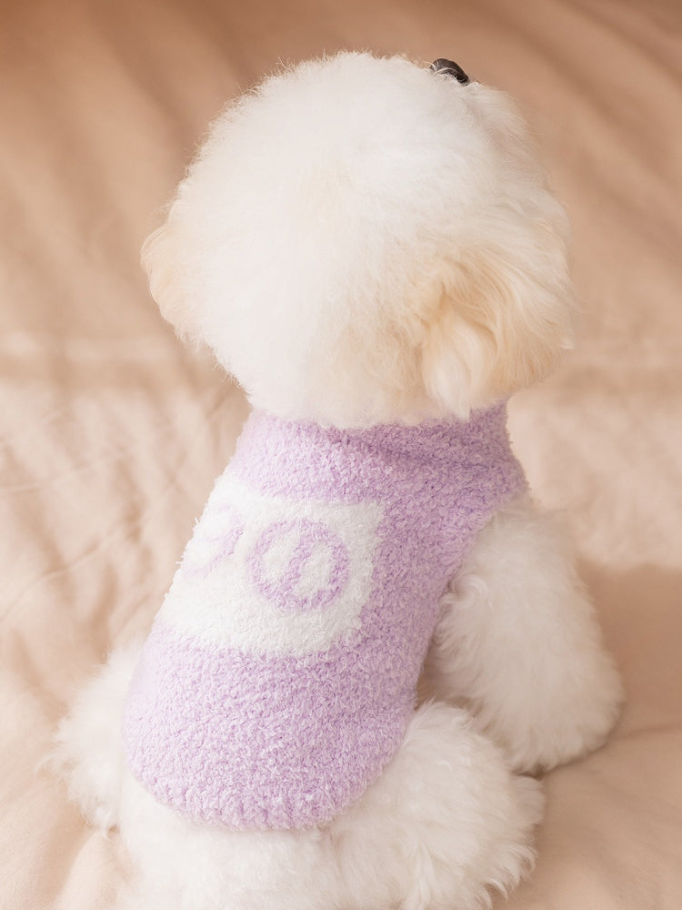 Rear view of a white bichon frise dog wearing a lavender fluffy dog sweater with a white graphic design on the back.