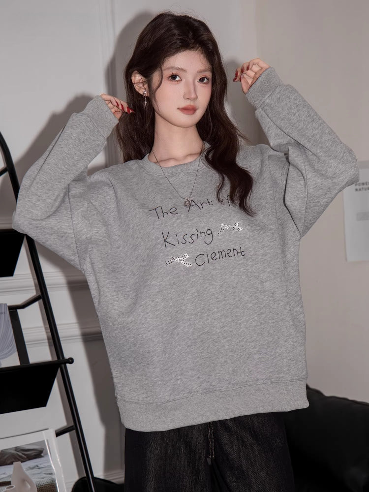 Woman wearing a grey long-sleeve sweatshirt with "The Art of Kissing Clement" graphic print.