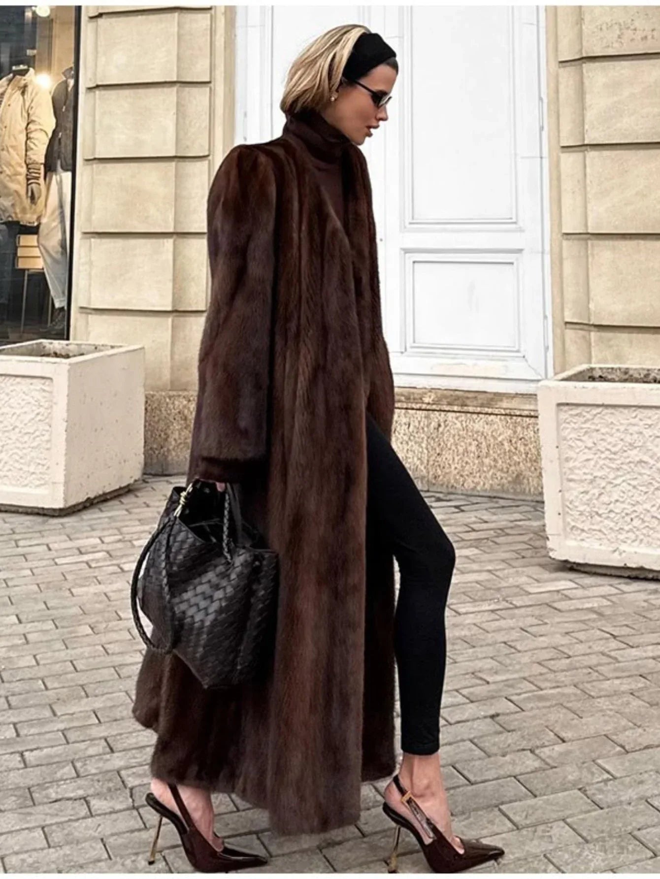 Woman wearing a long brown mink fur coat, black pants, and brown heels walking on a cobblestone street.