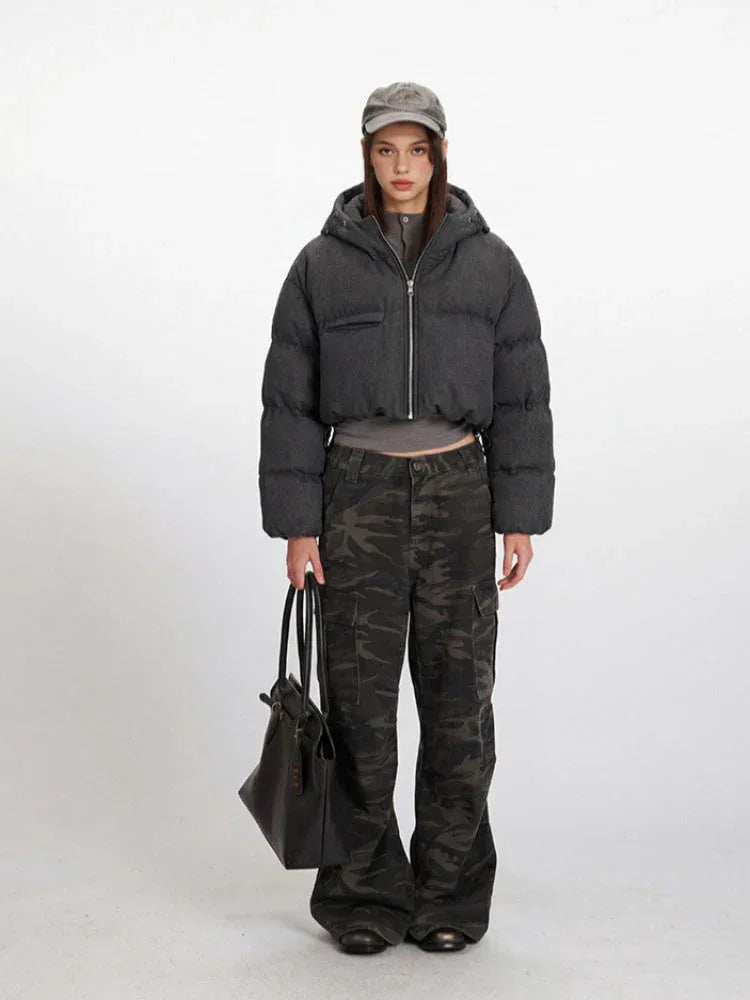 Woman wearing a cropped grey puffer jacket with a hood and zipper closure, paired with camouflage cargo pants and a grey baseball cap.