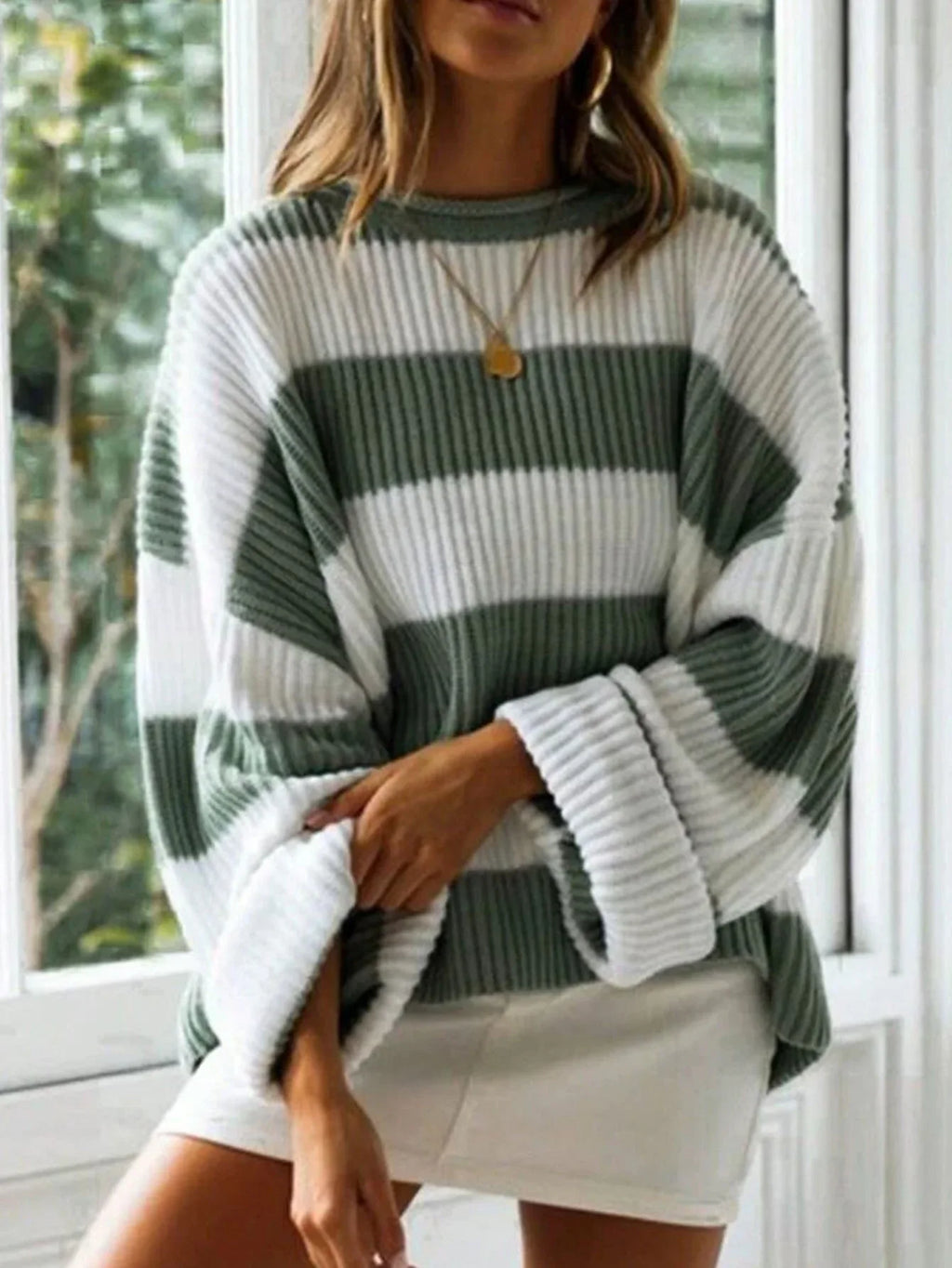 Woman in a green and white striped oversized knit sweater with crew neck and long cuffed sleeves, arm crossed.