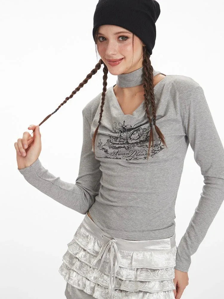 Model touching her braided hair, wearing a grey long-sleeve graphic t-shirt with a choker detail and silver ruffled shorts.