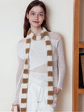 Woman posing with a brown and white striped fuzzy scarf around her neck, paired with a white top and white pants.