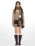 Woman wearing a beige V-neck knit sweater with long sleeves, paired with a brown patterned skirt, white socks, and brown boots.