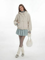 Model in a cream fuzzy hooded jacket with toggle closures, styled with a blue pleated skirt.