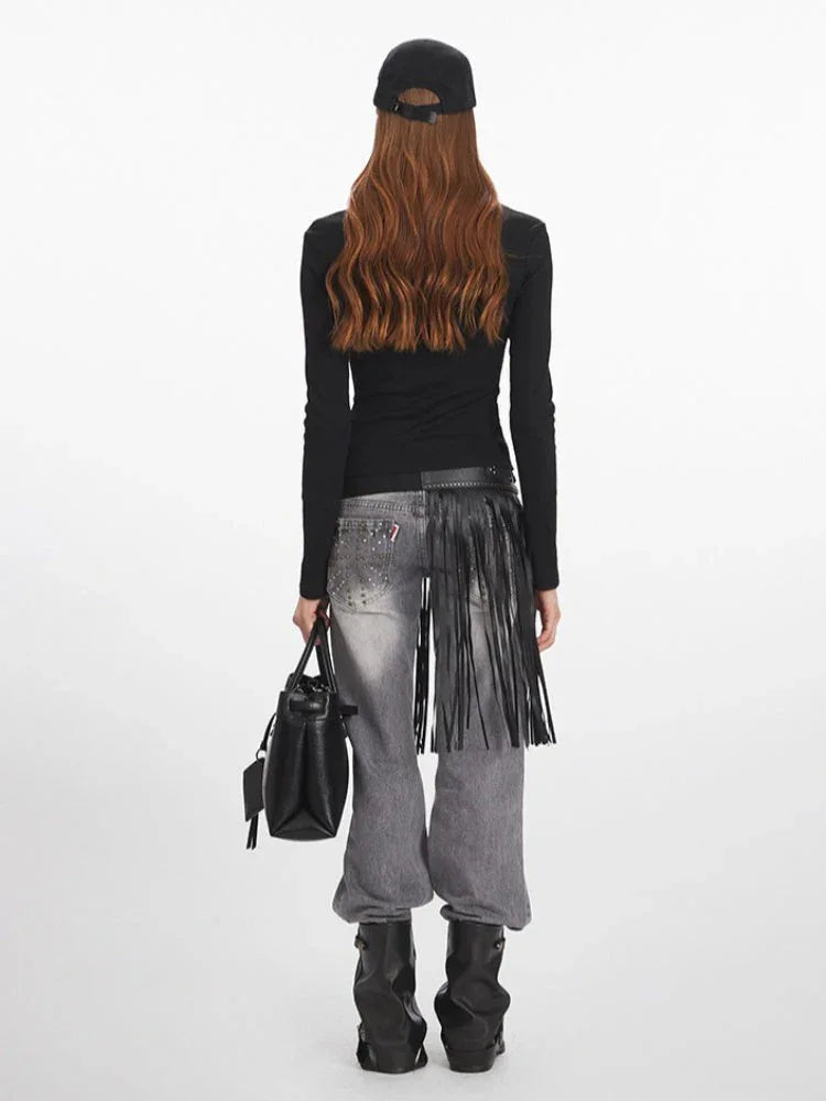 Back view of a woman wearing a black long-sleeve zip-up top with graphic print, grey wide-leg jeans with fringe detail, black boots, and black baseball cap.