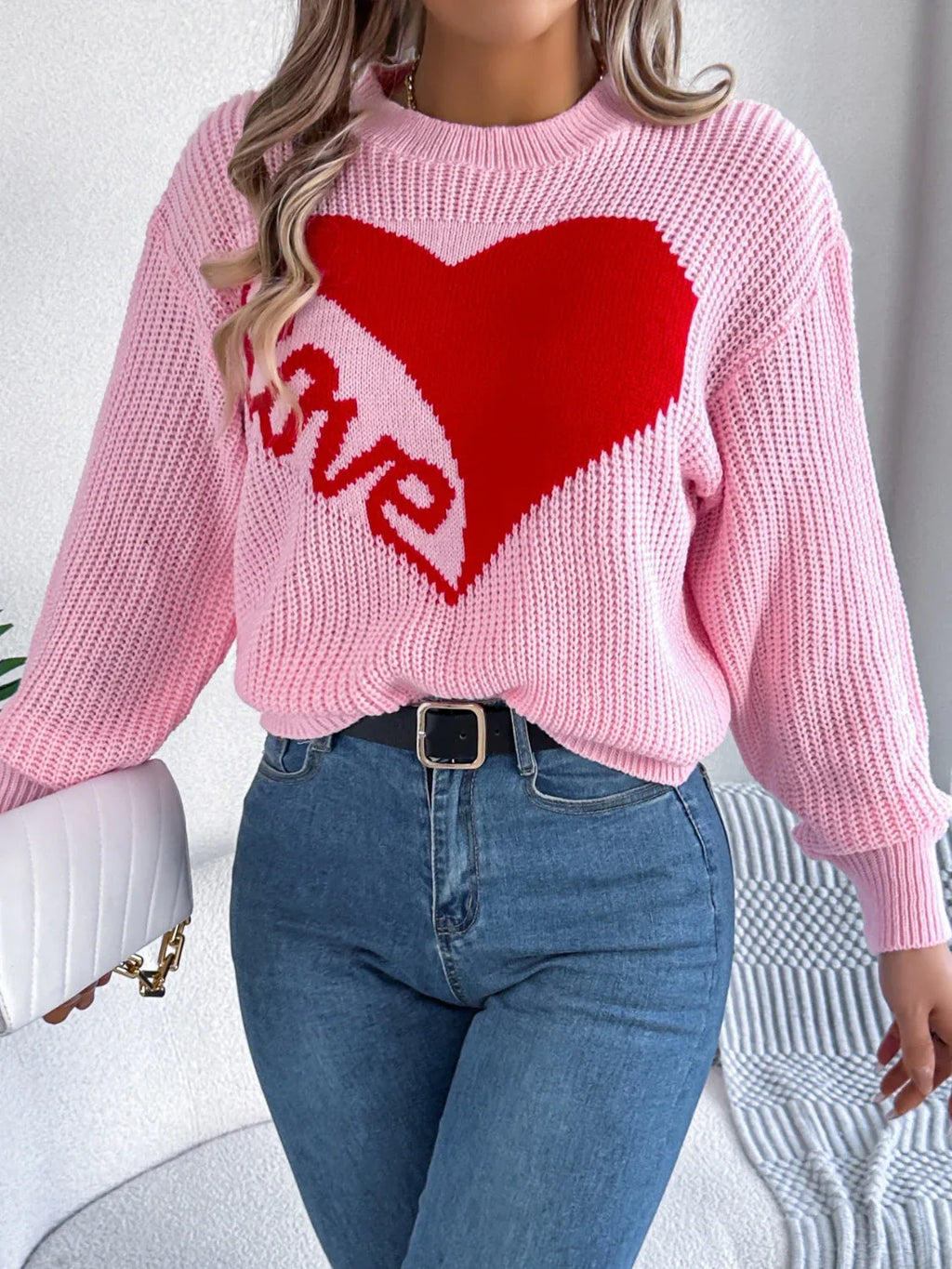 Woman holding a white handbag, showcasing a pink knit sweater with a heart design and blue jeans.