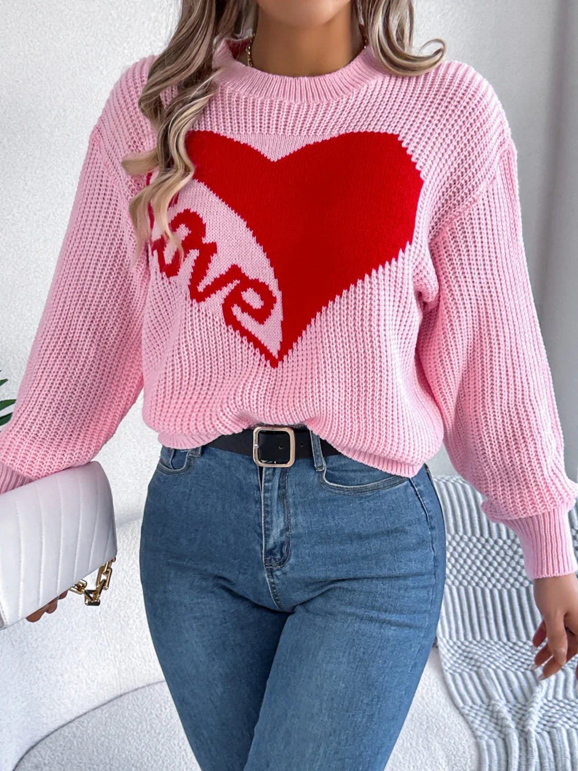 Woman holding a white handbag, showcasing a pink knit sweater with a heart design and blue jeans.