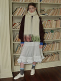 Full-length view of a woman with a cream scarf, maroon sweater, and white tiered skirt standing in front of bookshelves.