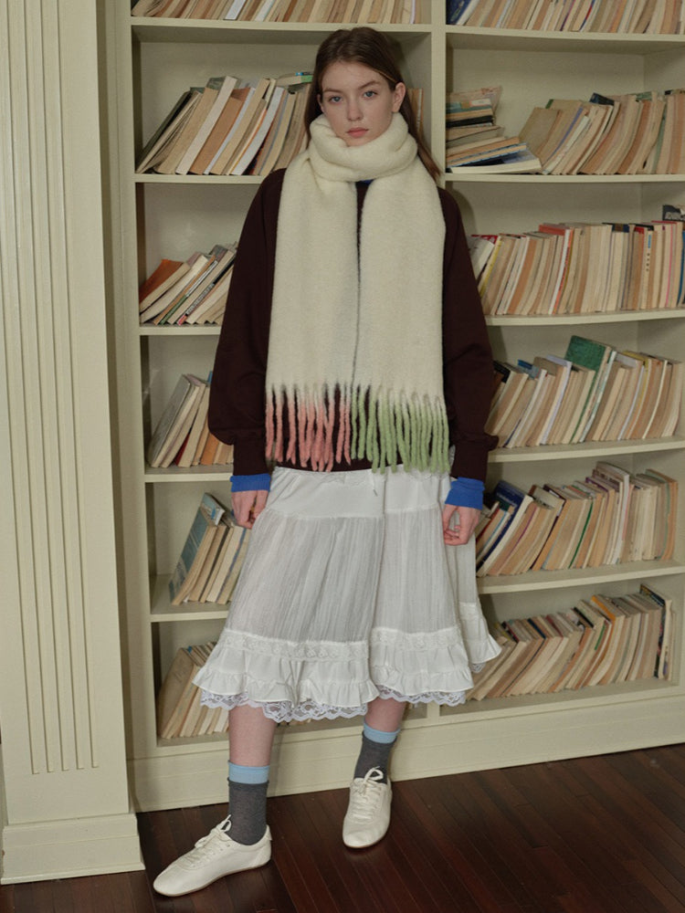 Full-length view of a woman with a cream scarf, maroon sweater, and white tiered skirt standing in front of bookshelves.