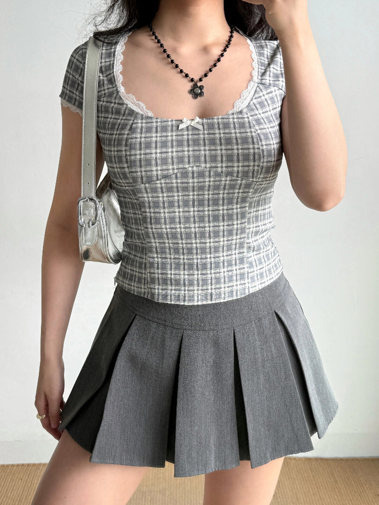 Front view of a woman in a grey plaid top and grey pleated skirt, holding a silver handbag.