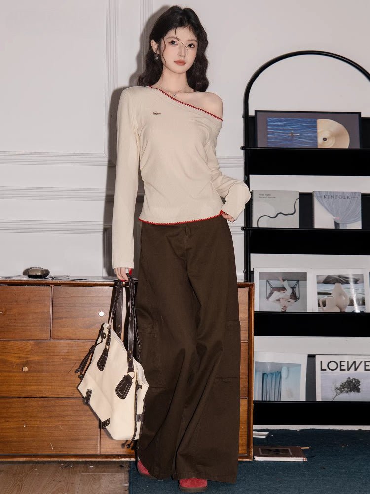 Full-body shot of a woman wearing a cream long-sleeve top with an asymmetrical neckline and red trim, dark brown cargo pants, and carrying a tote bag.