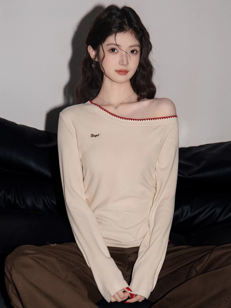 Woman wearing a cream long-sleeve top with an asymmetrical neckline and red trim, seated on a black couch.