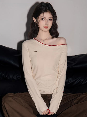 Woman wearing a cream long-sleeve top with an asymmetrical neckline and red trim, seated on a black couch.