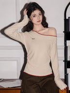 Woman wearing a cream long-sleeve top with an asymmetrical neckline and red trim, posing with her hand on her head.