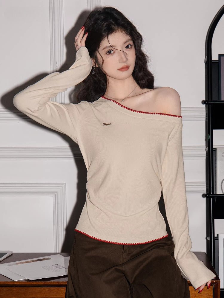 Woman wearing a cream long-sleeve top with an asymmetrical neckline and red trim, posing with her hand on her head.