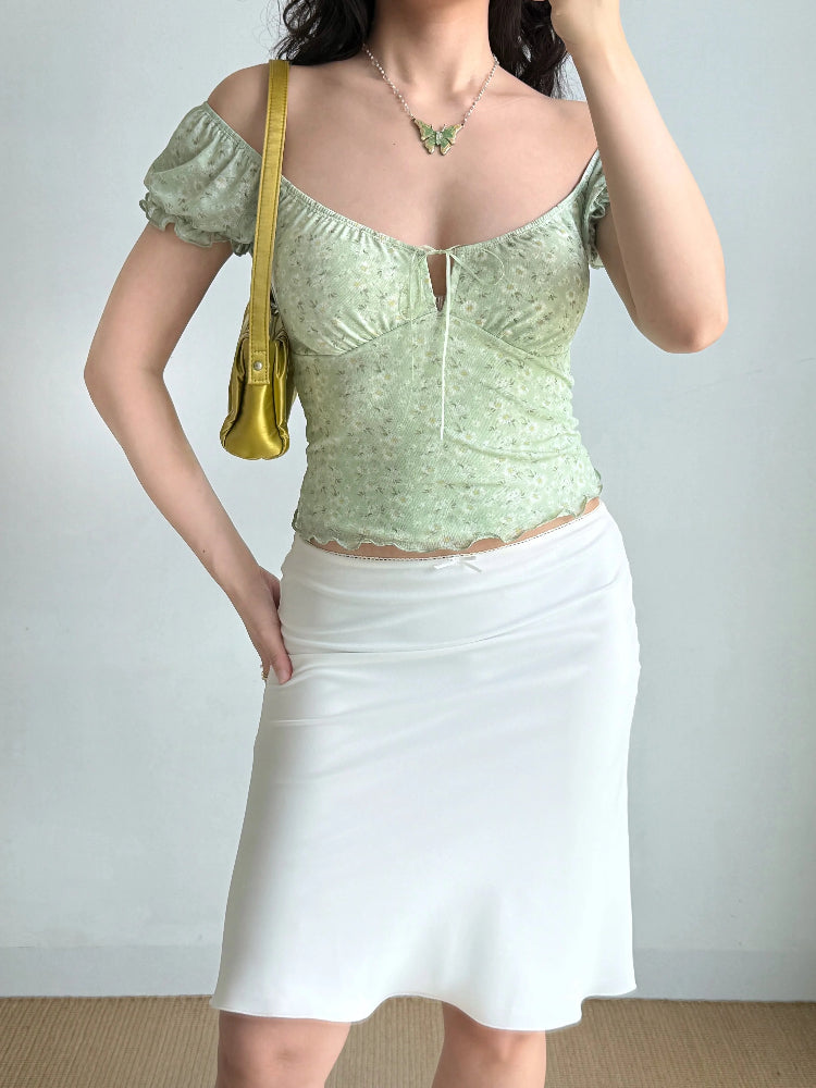 Full body shot of a woman wearing a green floral print top and a white skirt, accessorized with a gold handbag and butterfly necklace.