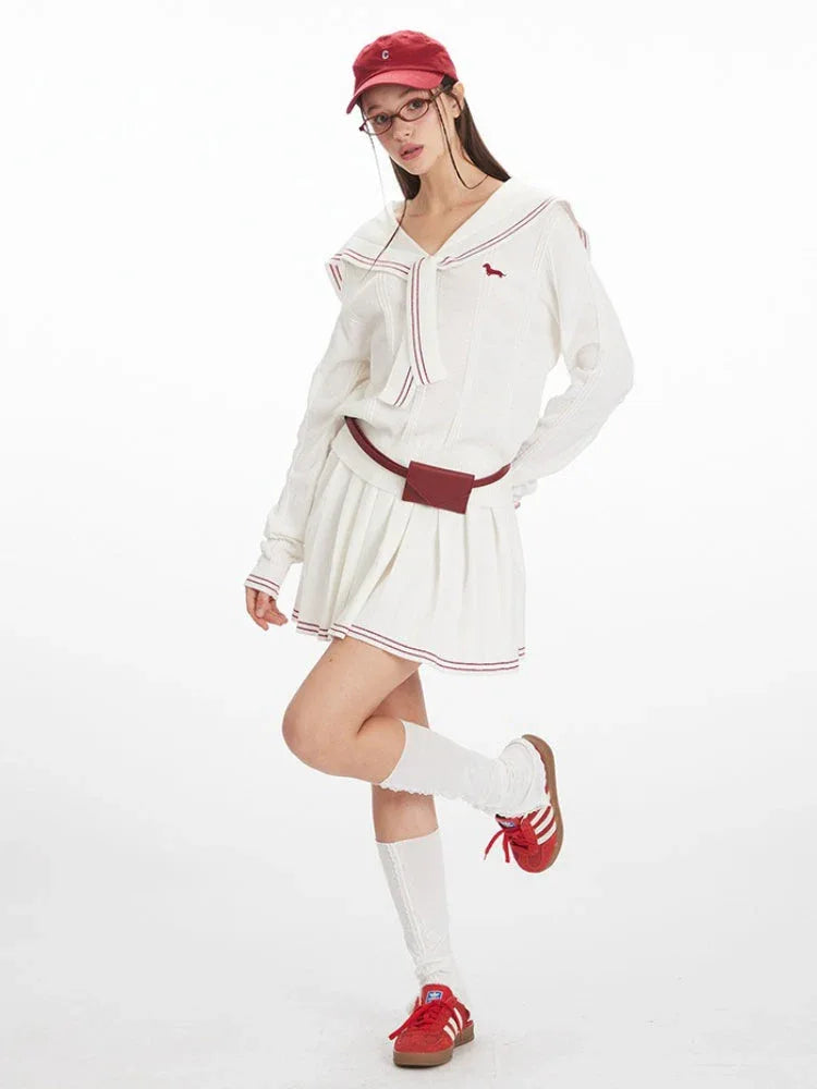 Full body shot of a woman wearing a white knit sweater with a navy sailor collar and red trim, a white pleated skirt, and red sneakers.