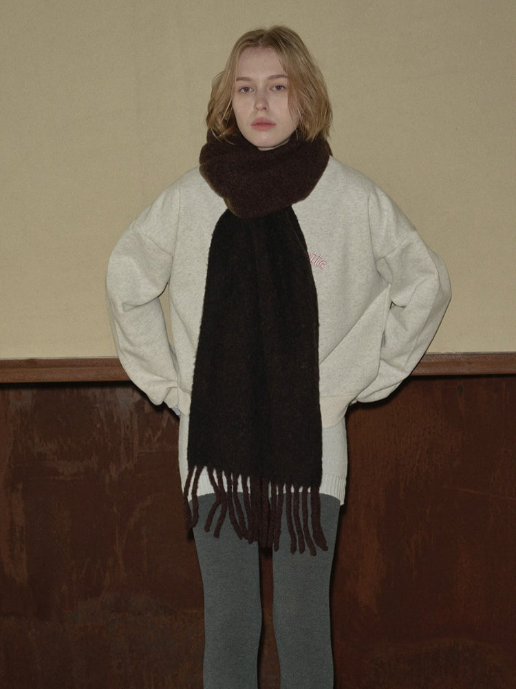 Woman wearing a light grey crewneck sweatshirt and a dark brown fringed scarf.