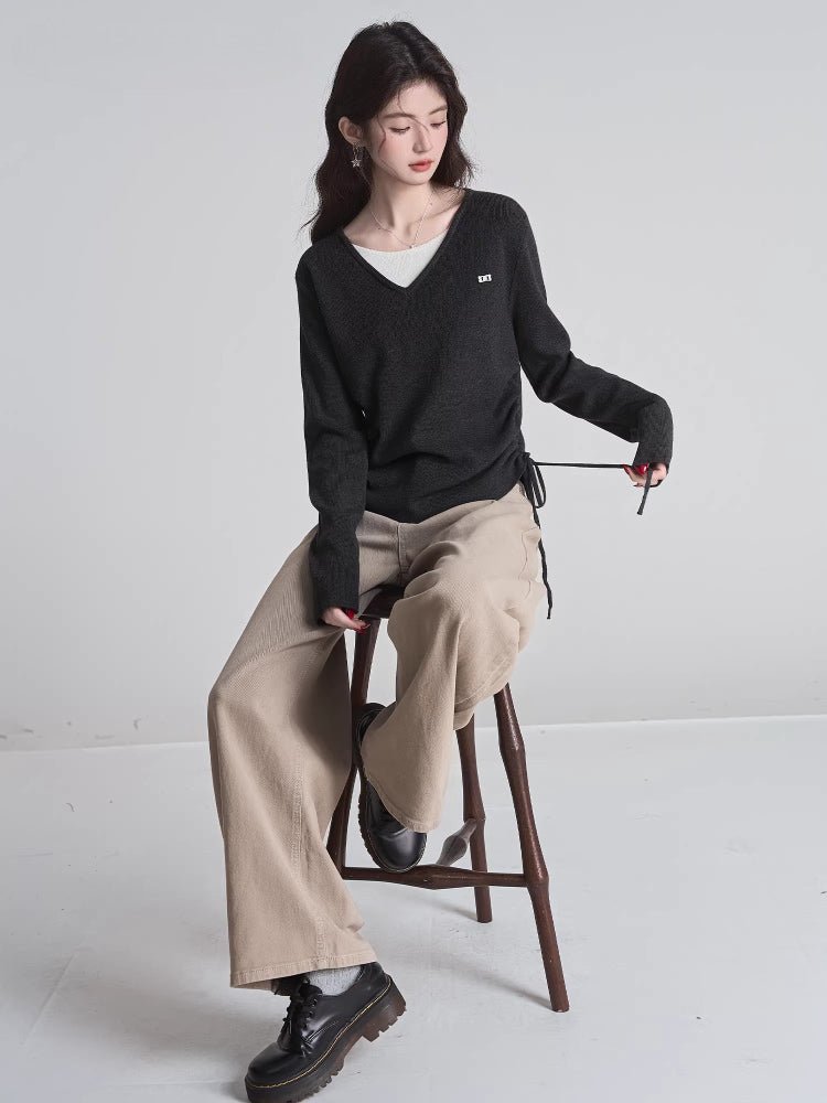 Full body shot of a woman seated on a stool, wearing a charcoal grey V-neck sweater, beige wide-leg pants, and black platform shoes.