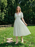 Woman wearing a light green and white striped midi dress with puff sleeves and a smocked waist, standing in a grassy field with trees in the background.