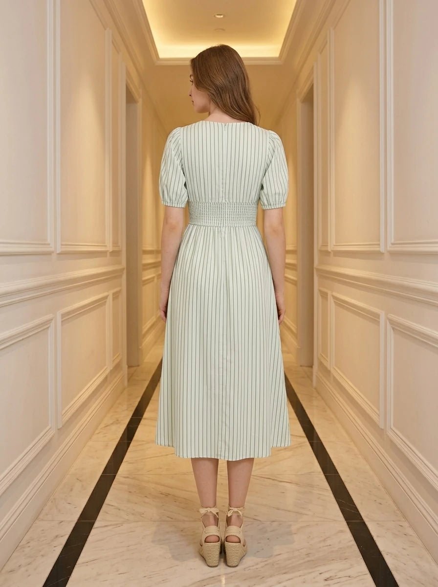 Woman wearing a light green and white striped midi dress with puff sleeves and a smocked waist, standing in a hallway.