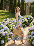 Woman wearing a fruit and floral print maxi dress, walking in a garden with hydrangeas, holding a straw bag.