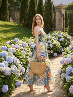 Woman wearing a fruit and floral print maxi dress, walking in a garden with hydrangeas, holding a straw bag.