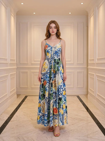 Woman wearing a full-length maxi dress with a colorful fruit and floral print, standing in a hallway.