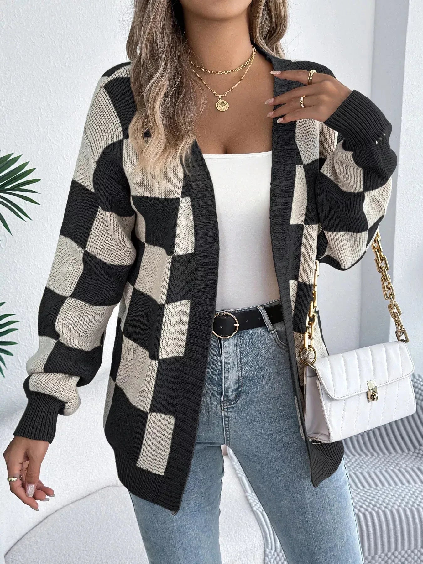 Woman wearing a black and cream checkerboard knit cardigan, white tank top, blue jeans, and gold jewelry.