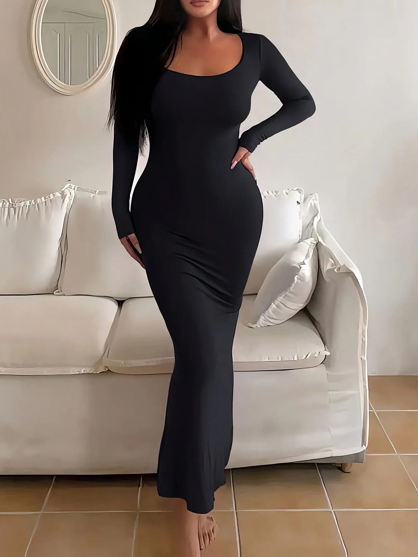 Woman in a long-sleeved, form-fitting, scoop neck black maxi dress standing in front of a white sofa.