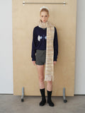 Model standing against a wooden backdrop wearing a navy argyle V-neck sweater, grey knit shorts, and a long striped scarf.
