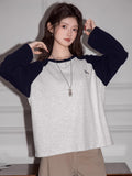 Woman adjusting her sleeves on a light grey long-sleeve raglan shirt with navy blue sleeves and a small embroidered bird graphic.