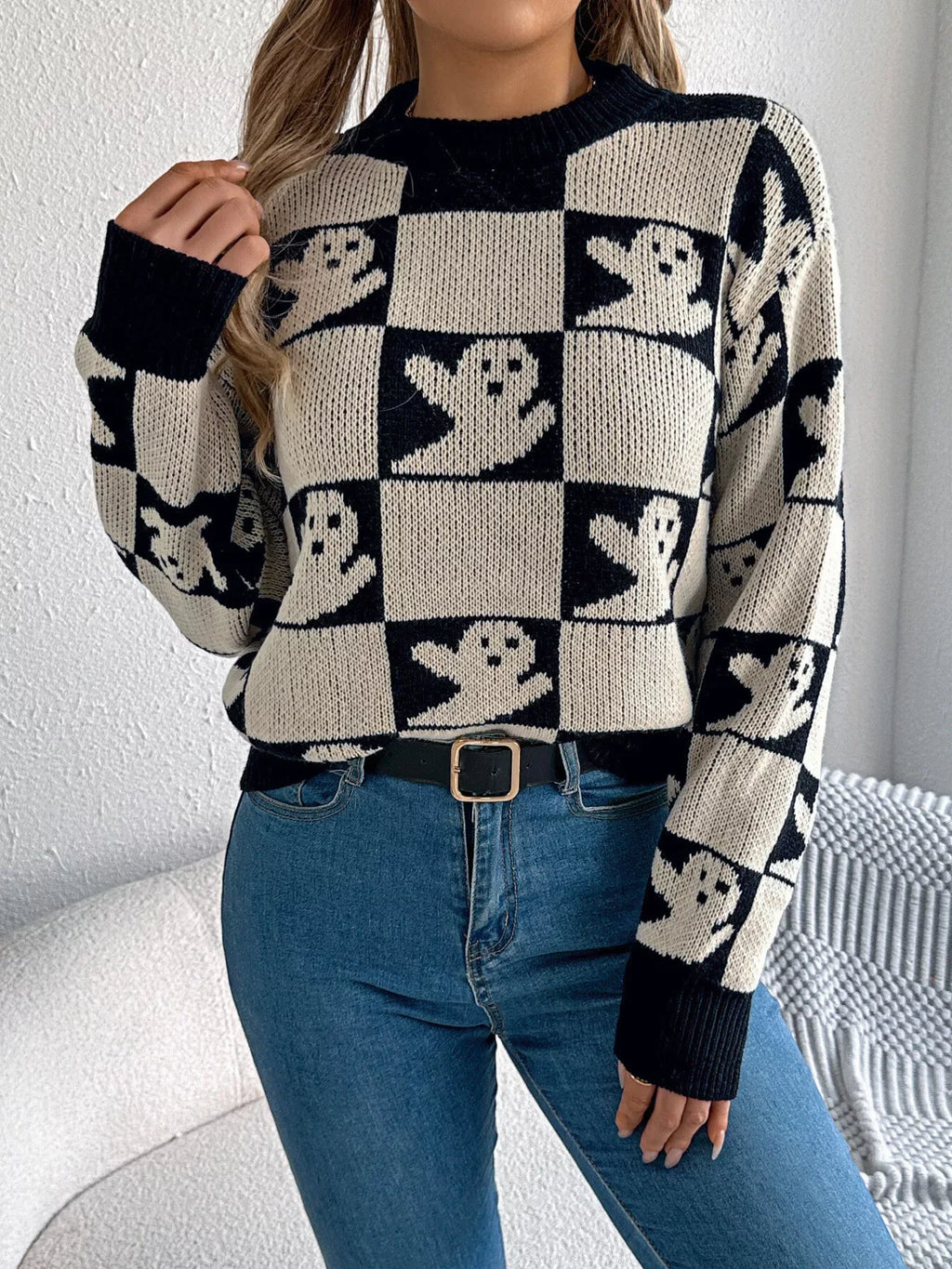 Front view of a woman wearing a black and cream checkerboard knit sweater with ghost print, blue jeans, and a black belt.
