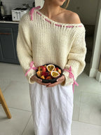 Woman holding a bowl of fruit, wearing a cream knit sweater with pink stitching and ties on the shoulders and sleeves.