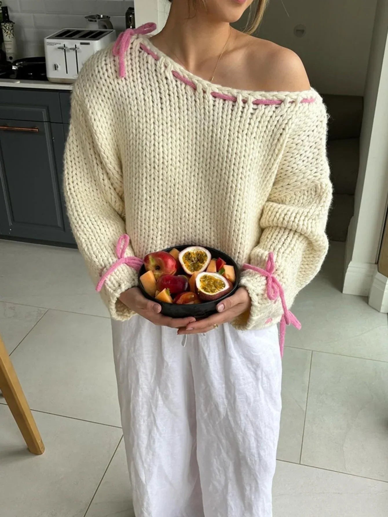 Woman holding a bowl of fruit, wearing a cream knit sweater with pink stitching and ties on the shoulders and sleeves.