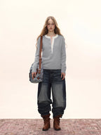 Woman wearing a light grey long-sleeved knit sweater with a white contrast neckline and button-up placket, carrying a denim crossbody bag and wearing distressed denim jeans and brown boots.
