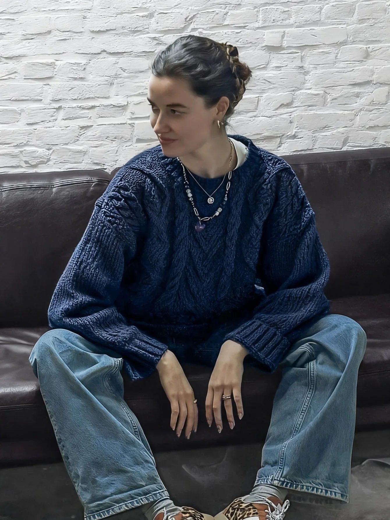 Woman sitting cross-legged on a brown leather couch, wearing a navy blue cable knit sweater and light wash jeans.
