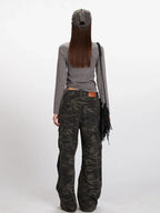 Back view of a model in a grey long-sleeve mock neck cropped top and camouflage cargo pants, with a grey baseball cap and a black fringed crossbody bag.