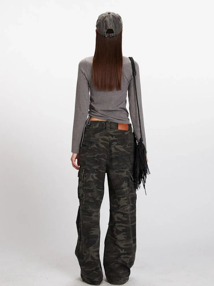 Back view of a model in a grey long-sleeve mock neck cropped top and camouflage cargo pants, with a grey baseball cap and a black fringed crossbody bag.