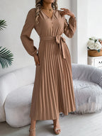 Woman in a brown long-sleeve V-neck midi dress with a pleated skirt and a self-tie belt, posing with hand on hip