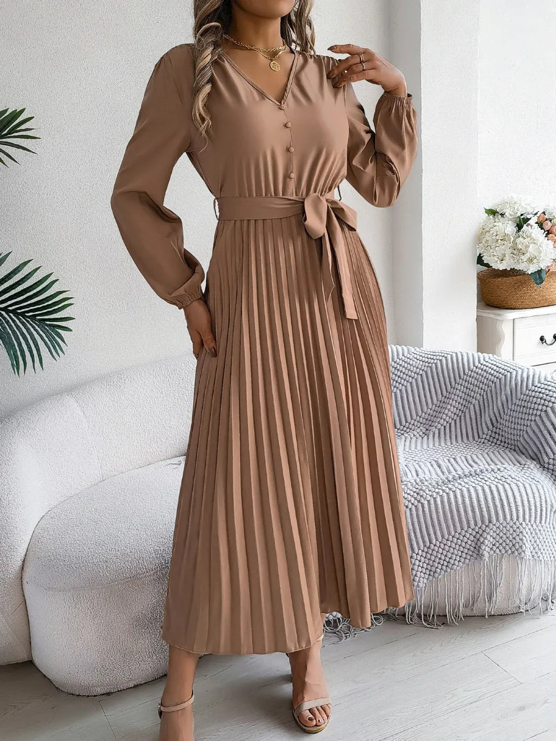 Woman in a brown long-sleeve V-neck midi dress with a pleated skirt and a self-tie belt, posing with hand on hip
