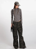 Full-body shot of a model wearing a casual outfit consisting of a grey long-sleeve mock neck cropped top, camouflage cargo pants, a grey baseball cap, and a black fringed crossbody bag.